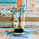 Sweet Summer silicone Apple Watch band with pastel pink, yellow, and blue surfboards, palm trees, and "sweet summer time" script on a soft watercolor beach scene, shown displayed and worn on Apple Watch against a coastal surfboard backdrop.