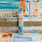 Sweet Summer silicone Samsung Galaxy Watch band with pastel pink, yellow, and blue surfboards, palm trees, and "sweet summer time" script on a soft watercolor beach scene, shown displayed and worn against a coastal surfboard backdrop.