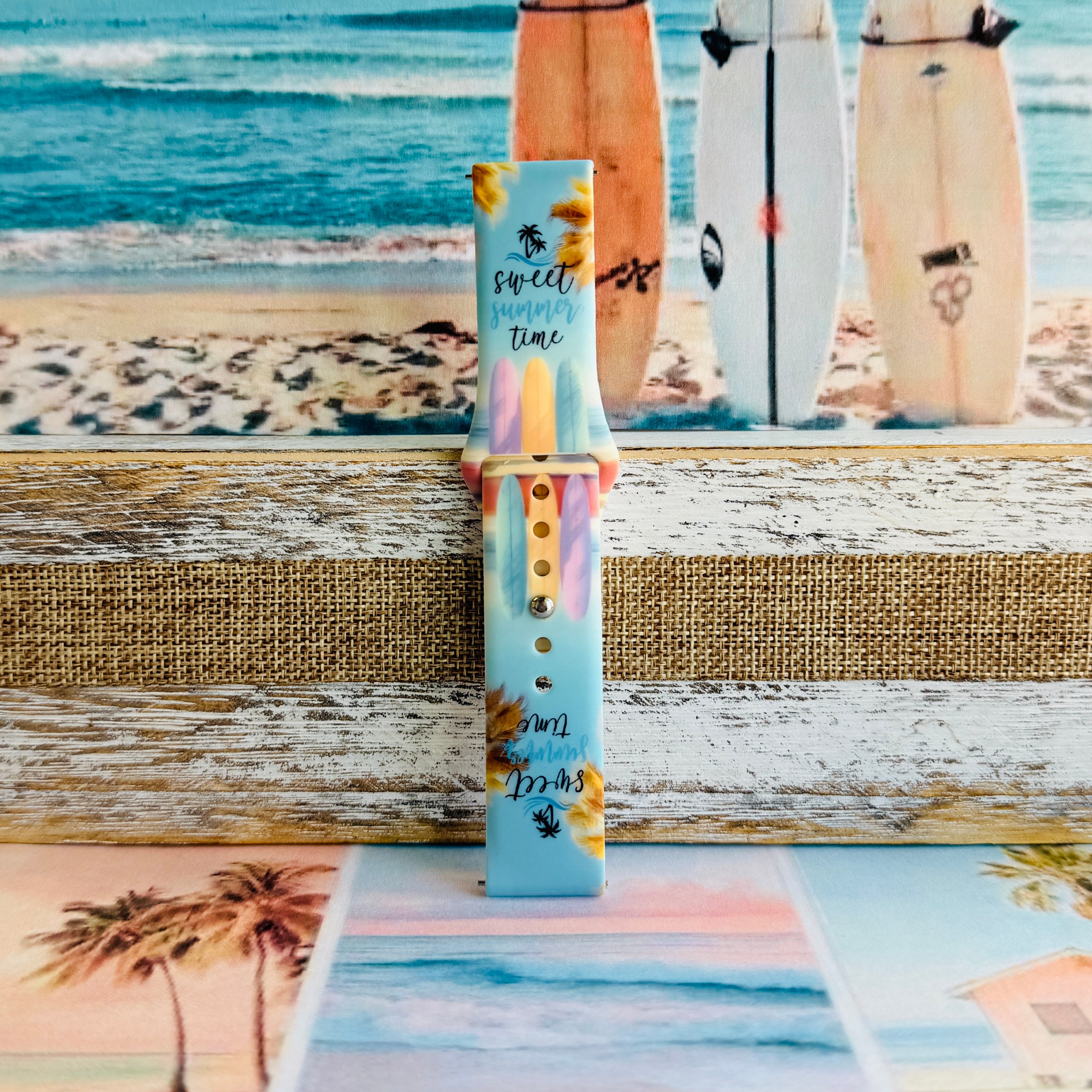 Sweet Summer silicone Samsung Galaxy Watch band with pastel pink, yellow, and blue surfboards, palm trees, and "sweet summer time" script on a soft watercolor beach scene, shown displayed and worn against a coastal surfboard backdrop.