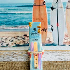 Sweet Summer silicone Samsung Galaxy Watch band with pastel pink, yellow, and blue surfboards, palm trees, and "sweet summer time" script on a soft watercolor beach scene, shown displayed and worn against a coastal surfboard backdrop.