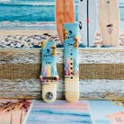 Sweet Summer silicone Samsung Galaxy Watch band with pastel pink, yellow, and blue surfboards, palm trees, and "sweet summer time" script on a soft watercolor beach scene, shown displayed and worn against a coastal surfboard backdrop.