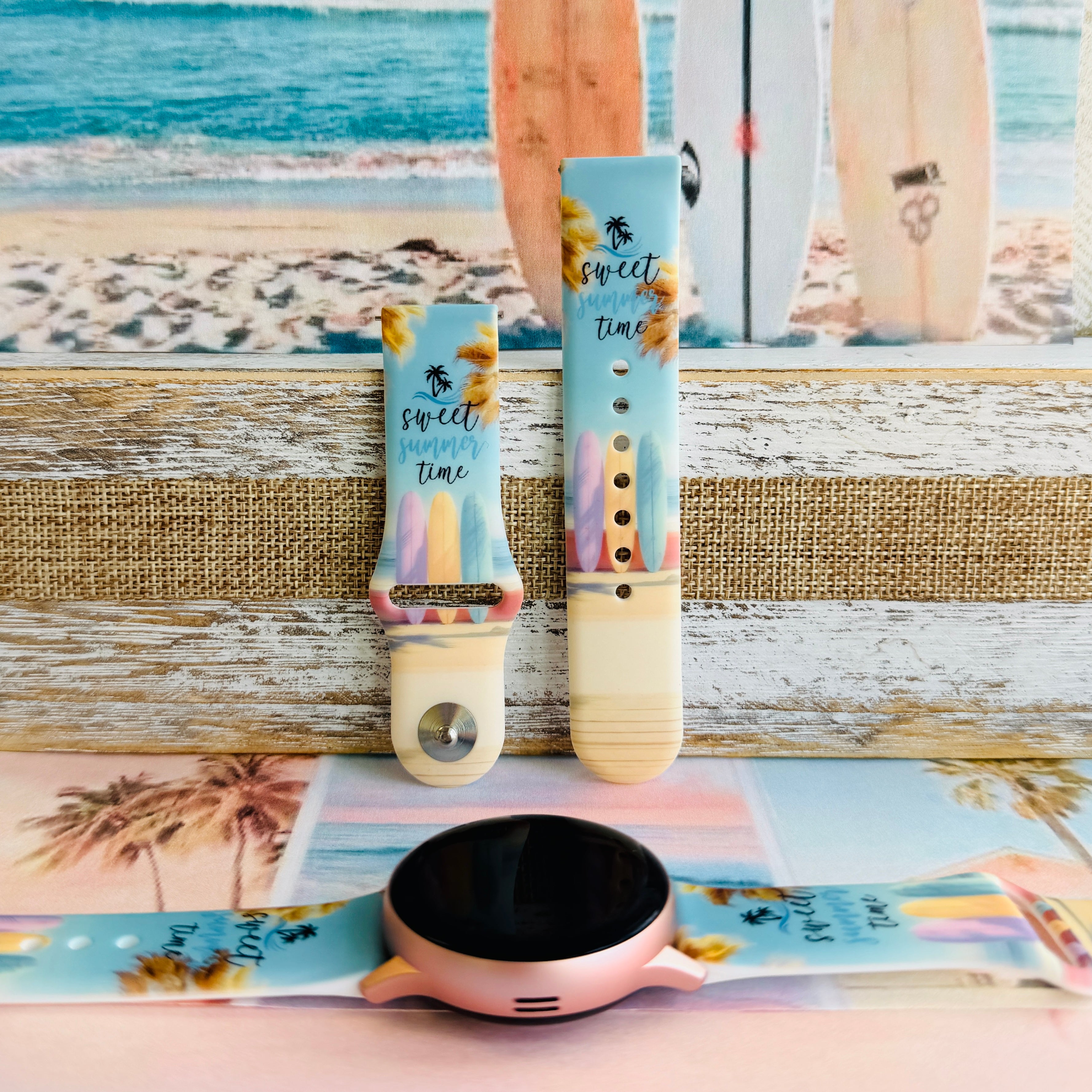 Sweet Summer silicone Samsung Galaxy Watch band with pastel pink, yellow, and blue surfboards, palm trees, and "sweet summer time" script on a soft watercolor beach scene, shown displayed and worn against a coastal surfboard backdrop.