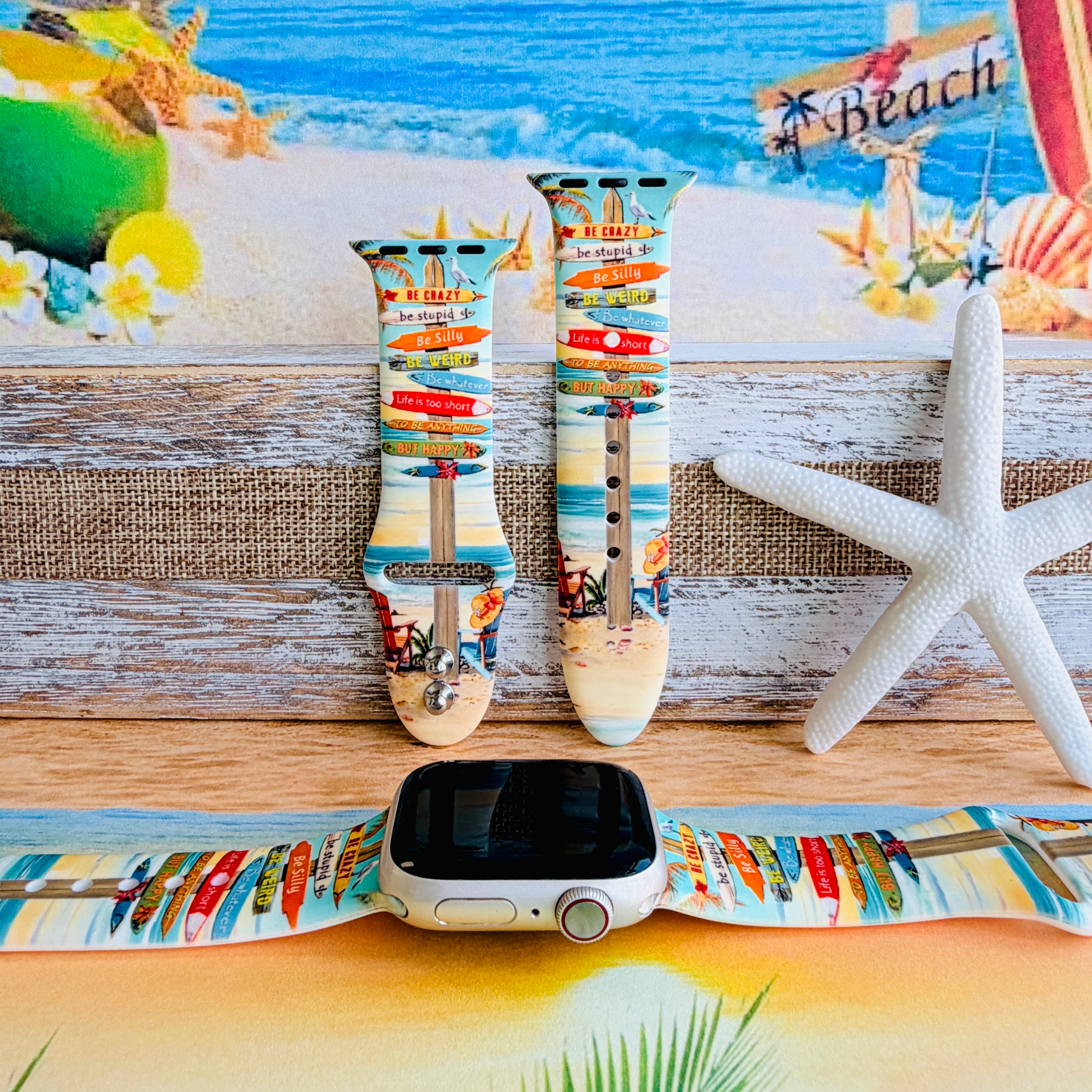 Seaside State of Mind silicone Apple Watch band with colorful surfboard signpost on a beach scene and "Be Crazy, Be Silly, Be Weird—life is too short to be anything but happy" message, shown displayed and worn on Apple Watch against a beach backdrop.