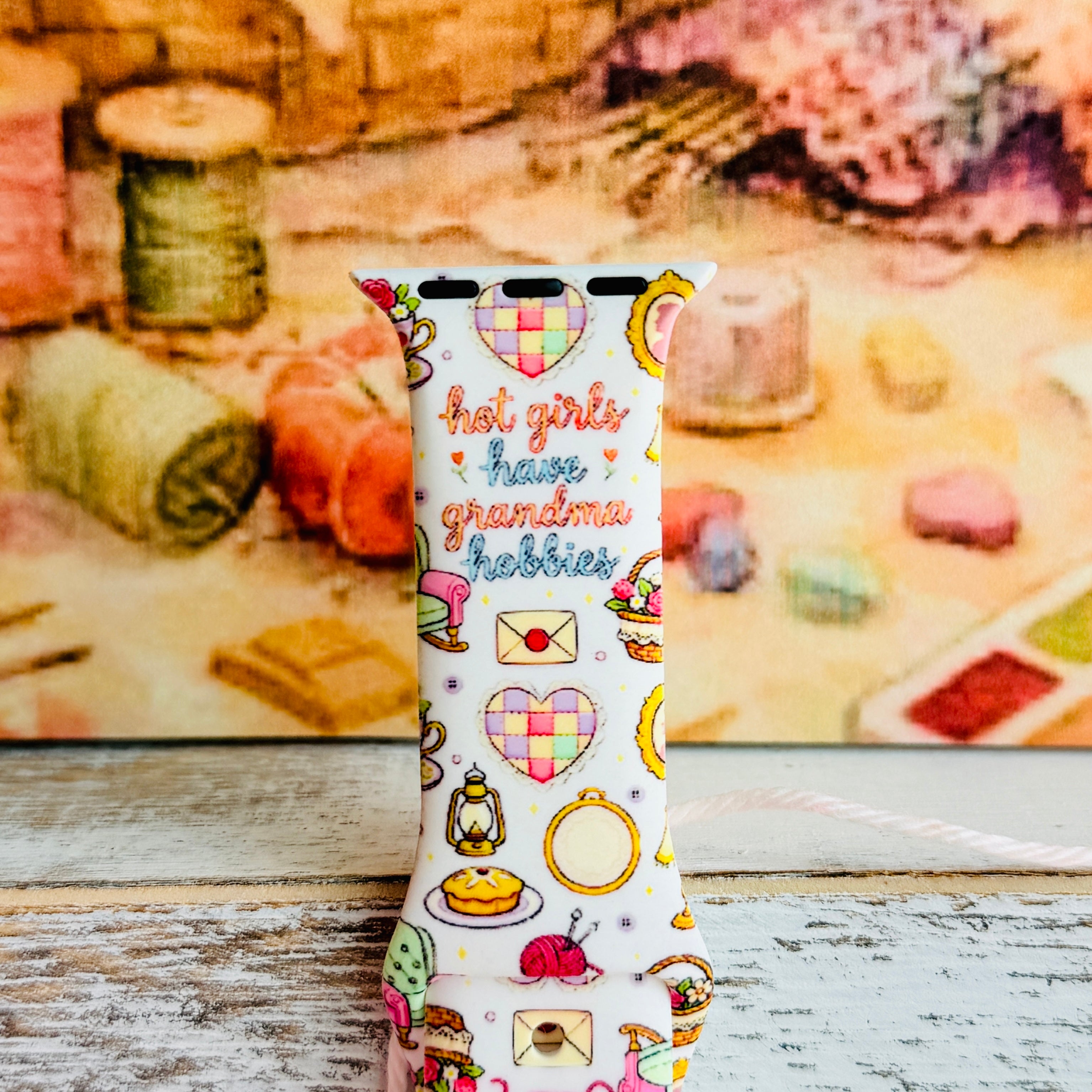 It's Giving Grandma Hobbies silicone Apple Watch band with "hot girls have grandma hobbies" script, patchwork hearts, yarn balls, knitting needles, embroidery hoops, lanterns, pies, and teacups on a cream base, shown displayed and worn on Apple Watch against a cottagecore craft backdrop.