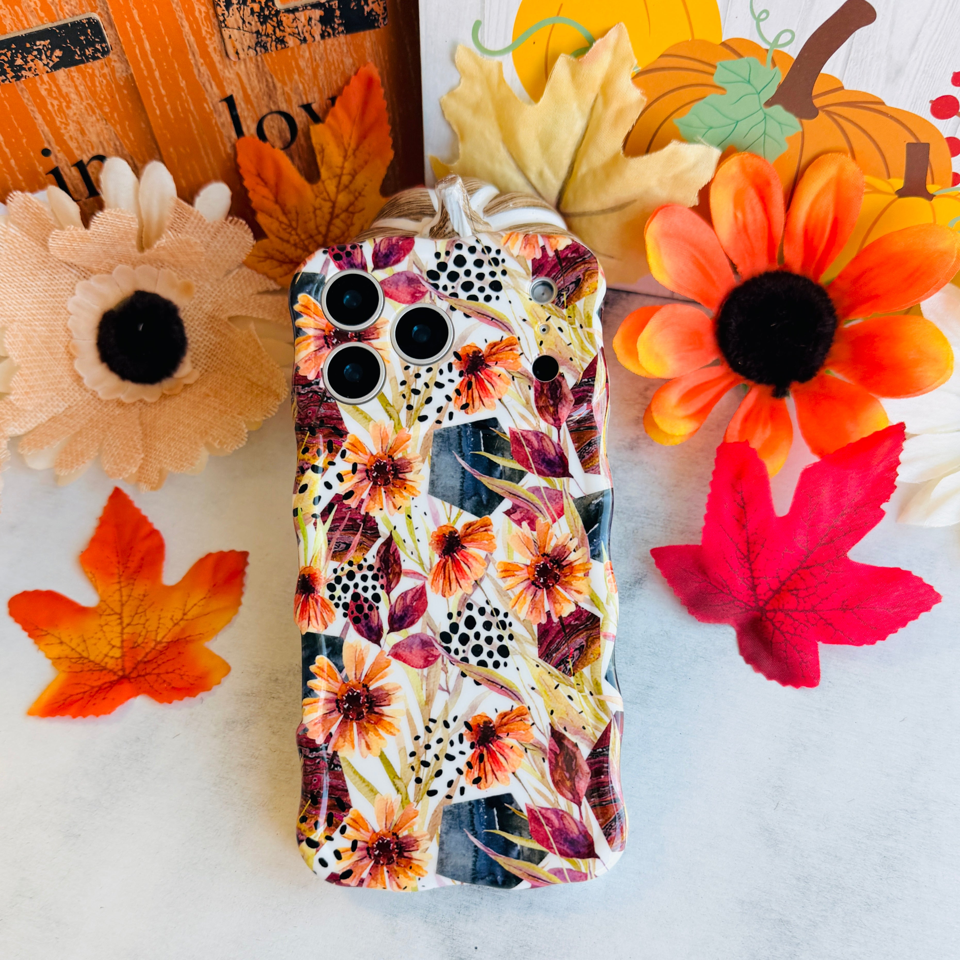 Sienna Petals Wavy Luxe MagSafe iPhone case laid flat among silk sunflowers and autumn maple leaves, showing the sculpted wavy edges and cream floral print.