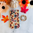Sienna Petals iPhone case beside its matching MagSafe grip ring on white marble with autumn maple leaves and a white knit pumpkin.