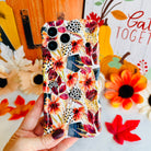 Hand holding the Sienna Petals Wavy Luxe MagSafe iPhone case in front of a Gather Together fall sign, cream with burnt orange watercolor flowers and black dot clusters.