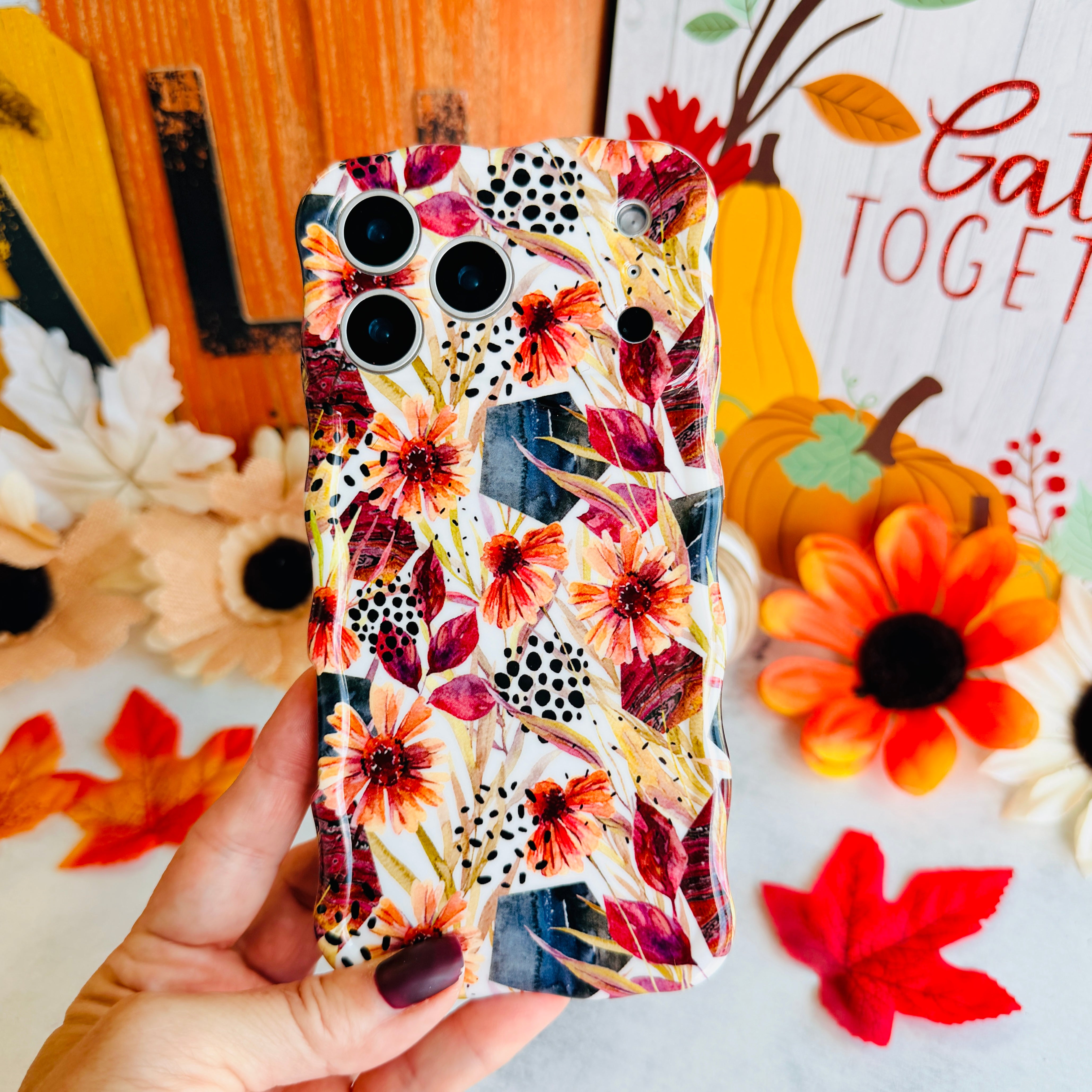 Hand holding the Sienna Petals Wavy Luxe MagSafe iPhone case in front of a Gather Together fall sign, cream with burnt orange watercolor flowers and black dot clusters.