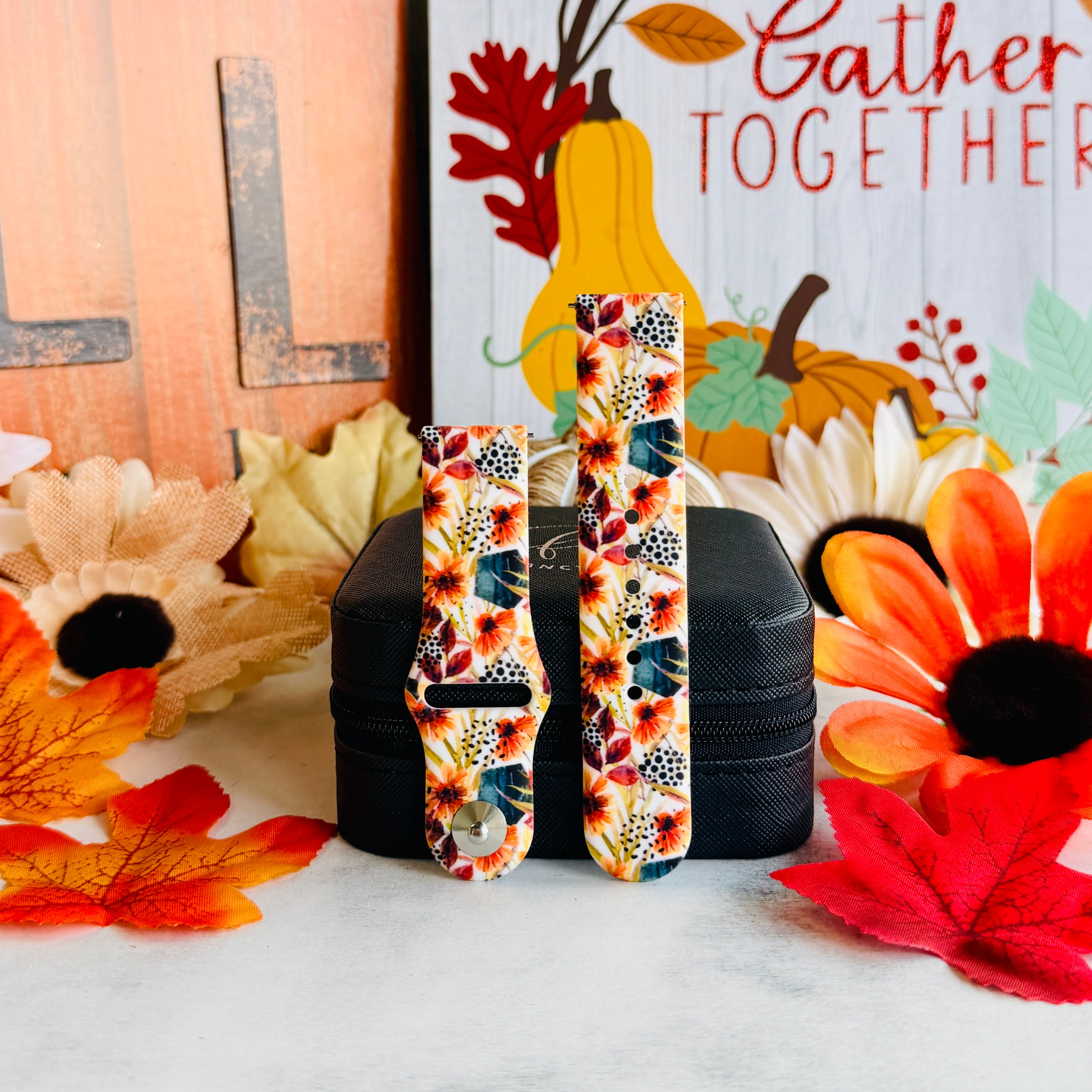 Sienna Petals Samsung Galaxy Watch band shown in both size options on a black stand against an autumn leaf and sunflower backdrop.