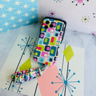 Mod Mood Scrunchie Phone Wrist Strap and matching Wavy Luxe phone case laid out together on a mid-century starburst print backdrop.