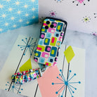 Mod Mood scrunchie phone wrist strap and matching Mod Mood Wavy Luxe iPhone case laid out together on a mid-century starburst backdrop