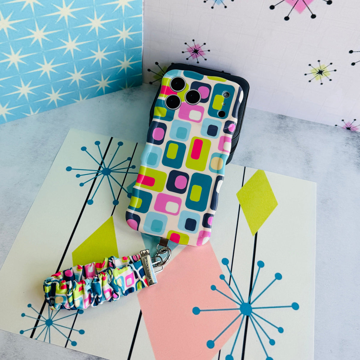 Mod Mood scrunchie phone wrist strap and matching Mod Mood Wavy Luxe iPhone case laid out together on a mid-century starburst backdrop