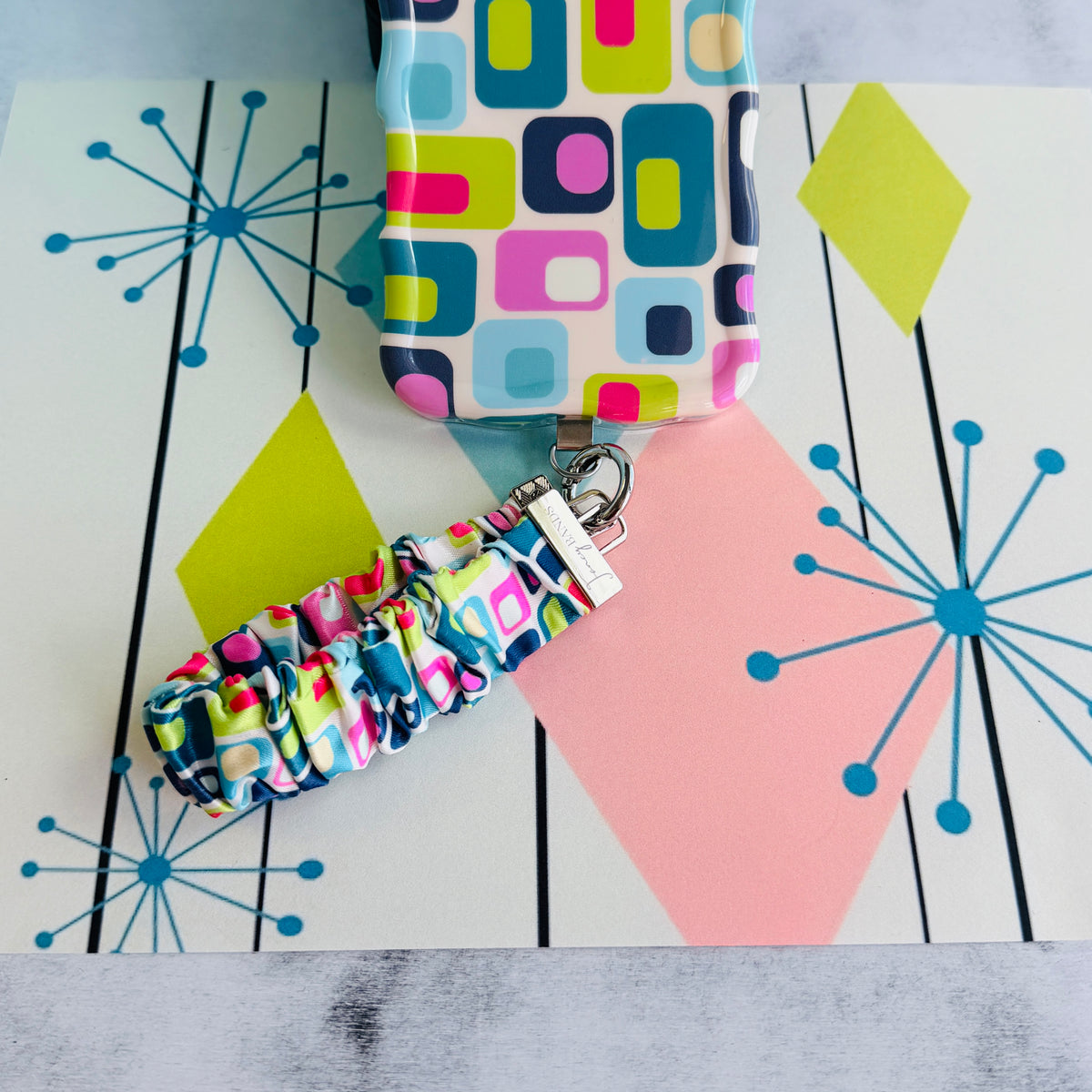 Mod Mood Scrunchie Phone Wrist Strap clipped to the matching retro geometric phone case, on an atomic starburst backdrop.