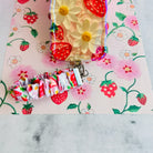 Berry Cute Blooms Scrunchie Phone Wrist Strap beside the matching strawberry and daisy phone case, on strawberry print paper.