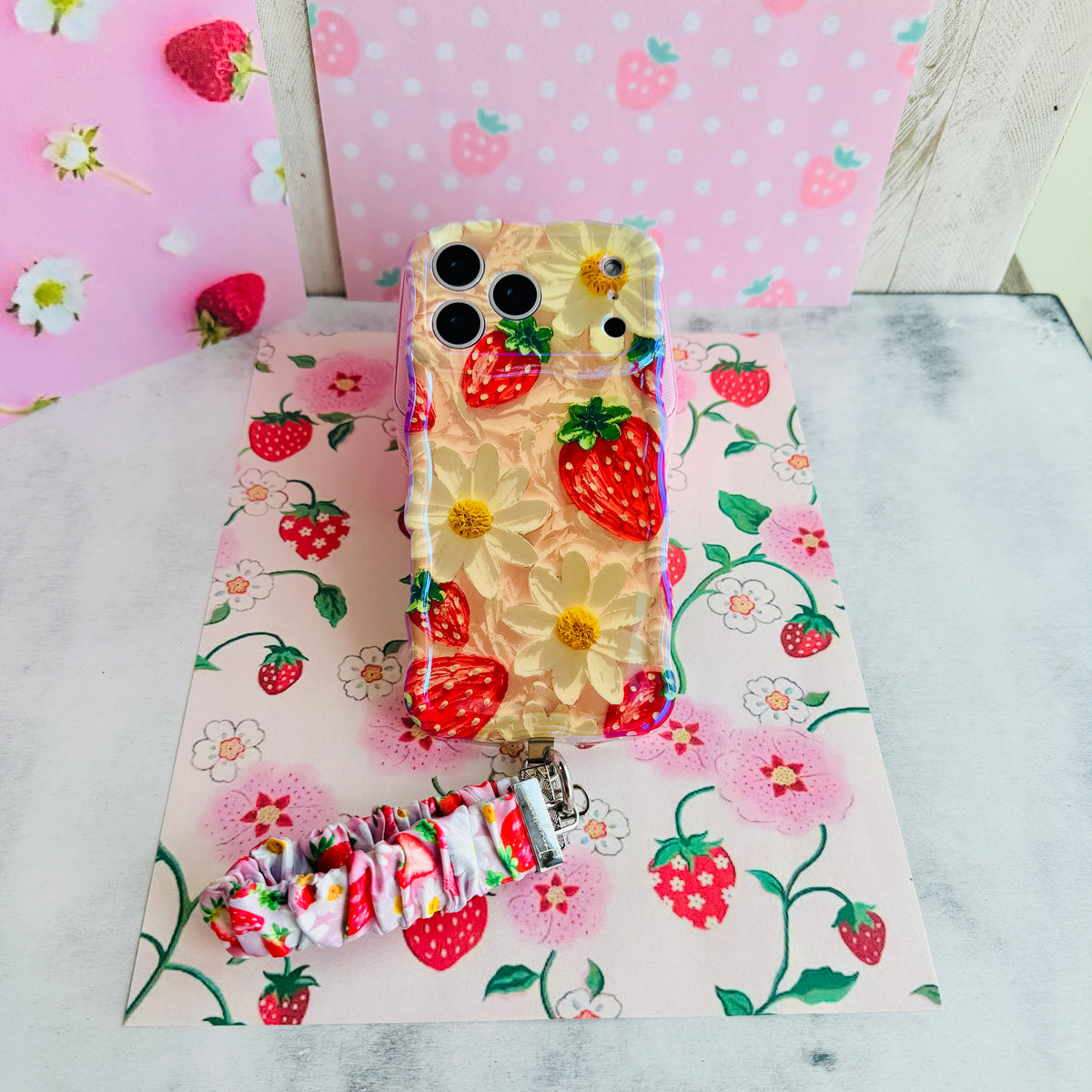 Berry Cute Blooms Wavy Luxe phone case standing with the matching scrunchie wrist strap, against a strawberry and daisy backdrop.