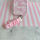 Blush Manor Scrunchie Phone Wrist Strap beside the matching pink toile phone case, on pink and white striped paper.