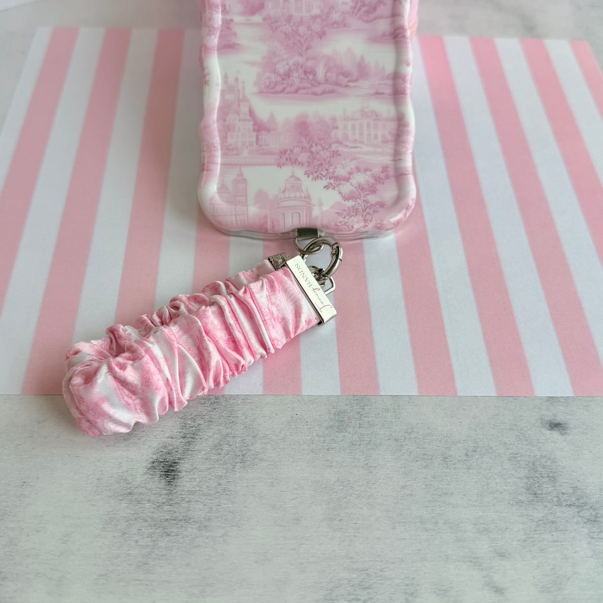 Blush Manor Scrunchie Phone Wrist Strap beside the matching pink toile phone case, on pink and white striped paper.