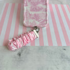 Blush Manor scrunchie phone wrist strap clipped to the matching pink toile Wavy Luxe iPhone case, on pink and white striped paper