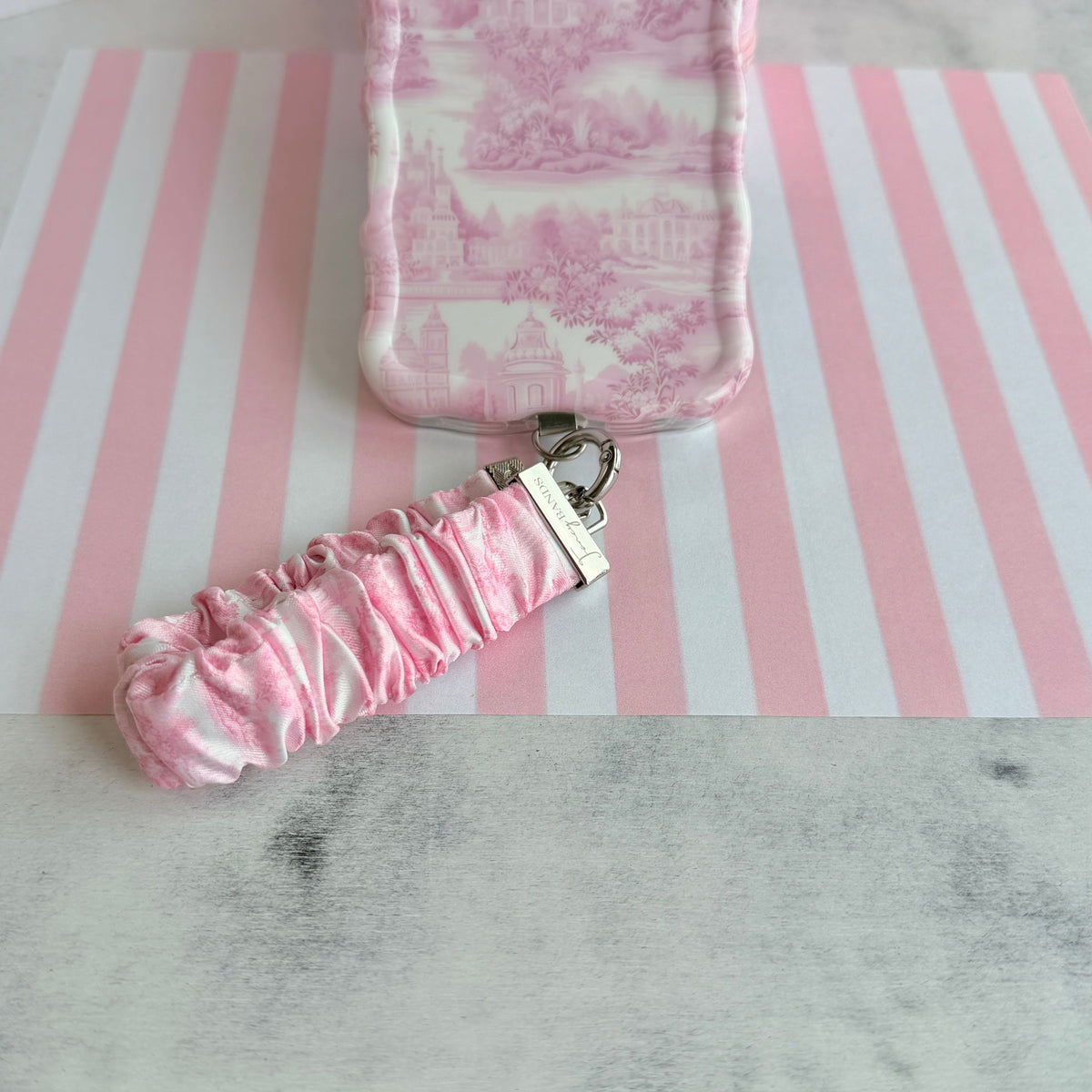 Blush Manor scrunchie phone wrist strap clipped to the matching pink toile Wavy Luxe iPhone case, on pink and white striped paper