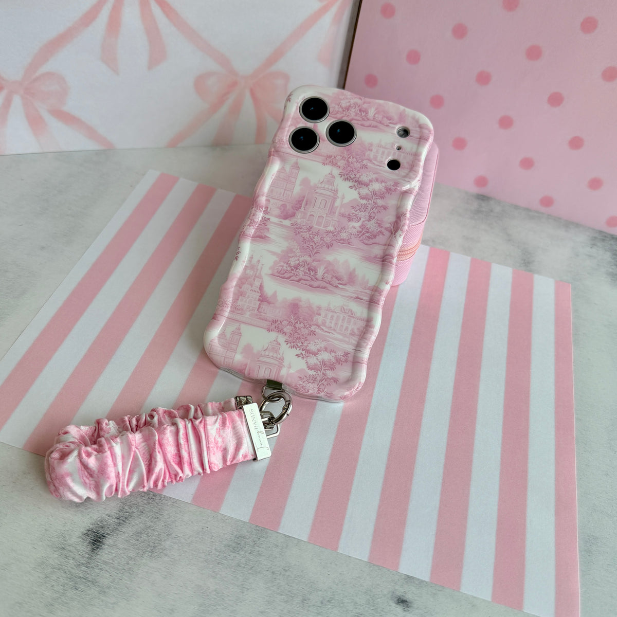 Blush Manor Wavy Luxe phone case standing with the matching pink toile scrunchie wrist strap, against a pink stripe and bow backdrop.