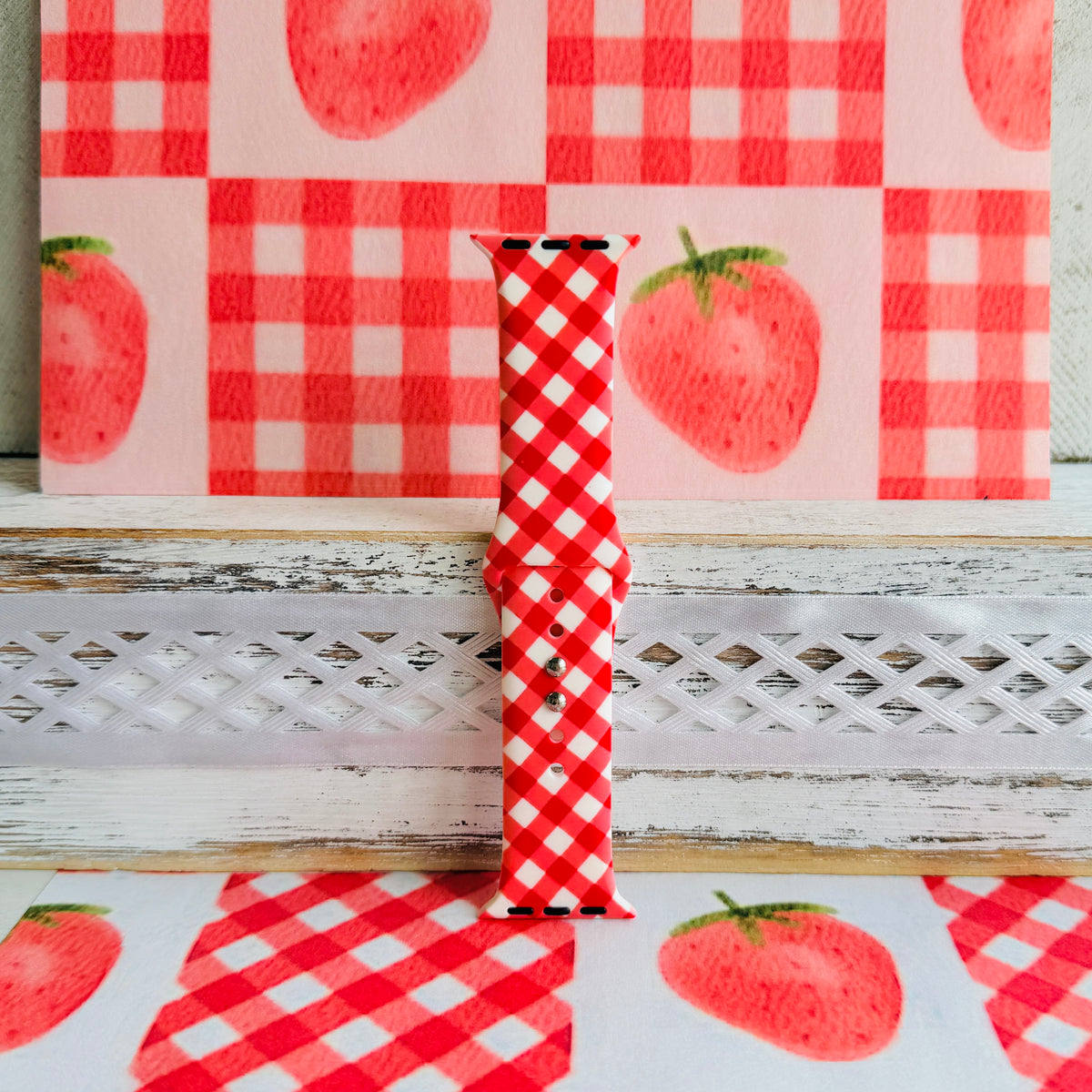Red Barn Babe silicone Apple Watch band standing full length on a red gingham picnic cloth with painted strawberries.