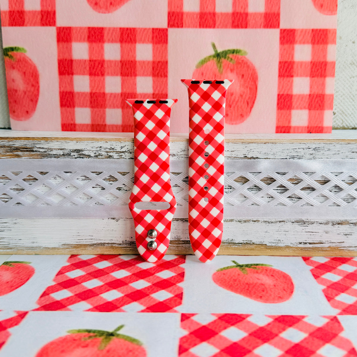 Red Barn Babe Apple Watch band in both size options on a whitewashed wood ledge, red gingham print against a strawberry backdrop.