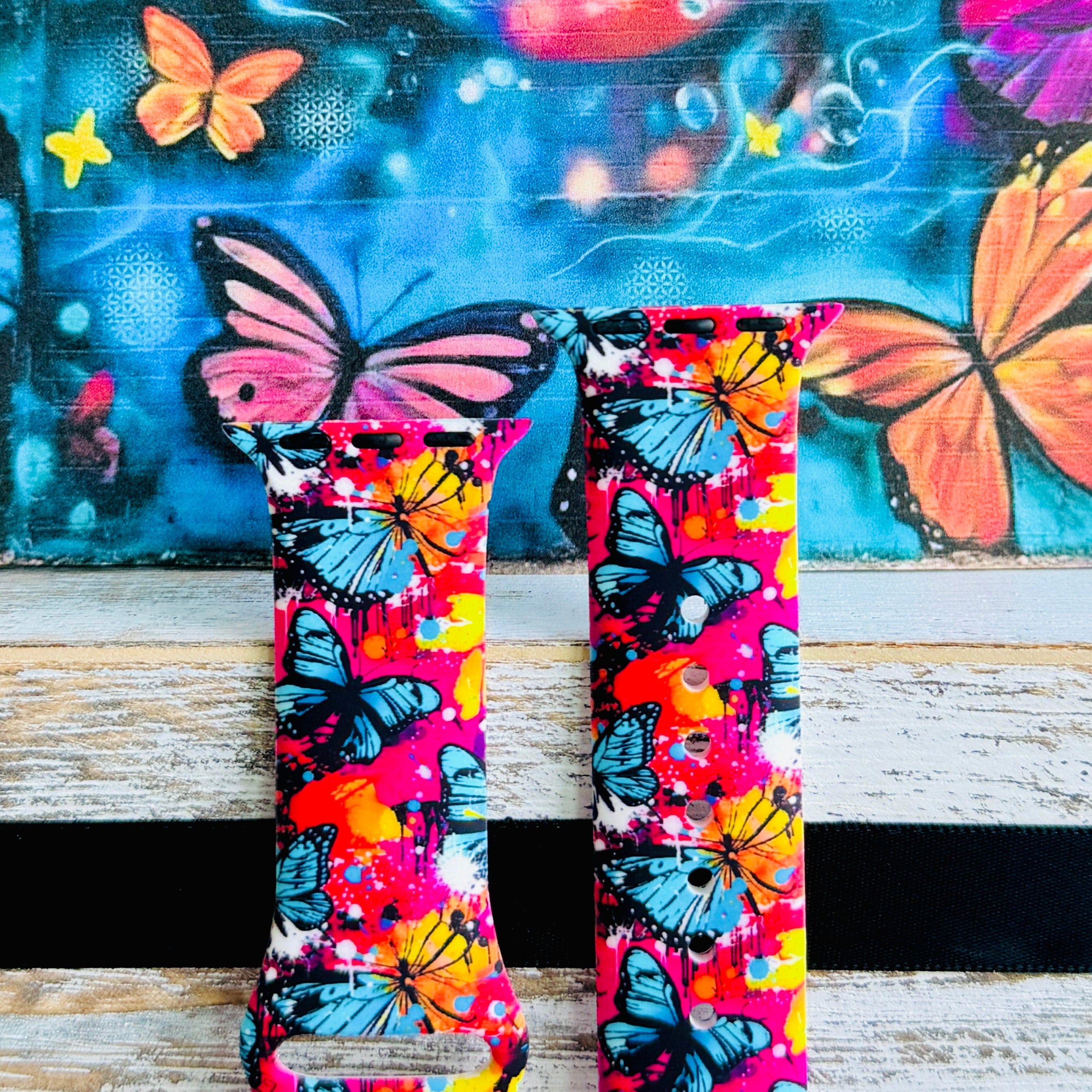 Rebel Wings silicone Apple Watch band with graffiti-style butterflies in blue, orange, yellow, and white over a hot pink and magenta paint-splatter background, shown displayed and worn on Apple Watch against a colorful street art butterfly backdrop.