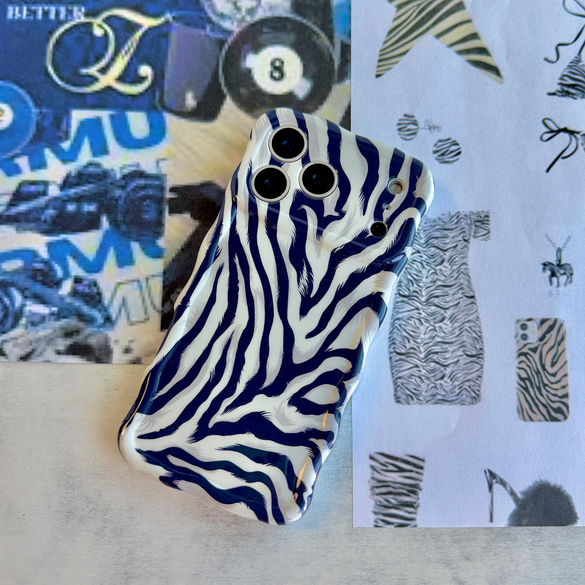 The Zebra, Obviously navy and white zebra print MagSafe iPhone case propped on a blue fashion magazine collage, showing the sculpted wavy border and the full stripe pattern.
