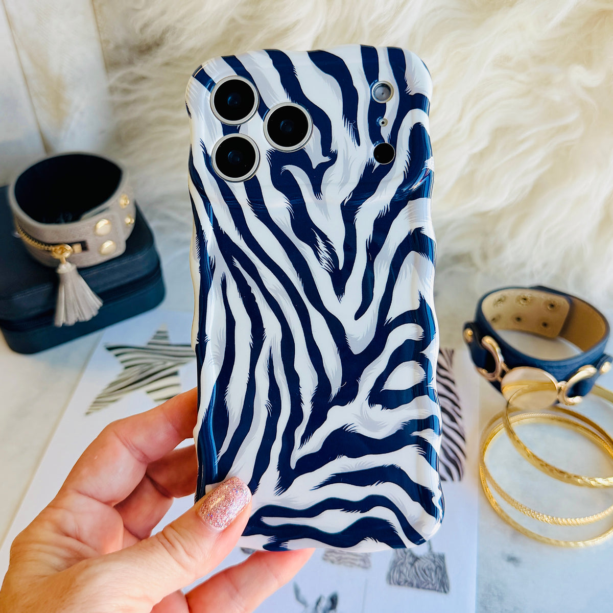 Hand holding the Zebra, Obviously navy and white zebra print Wavy Luxe iPhone case over a fashion flat lay with gold bangles, a studded cuff and animal-print magazine clippings.