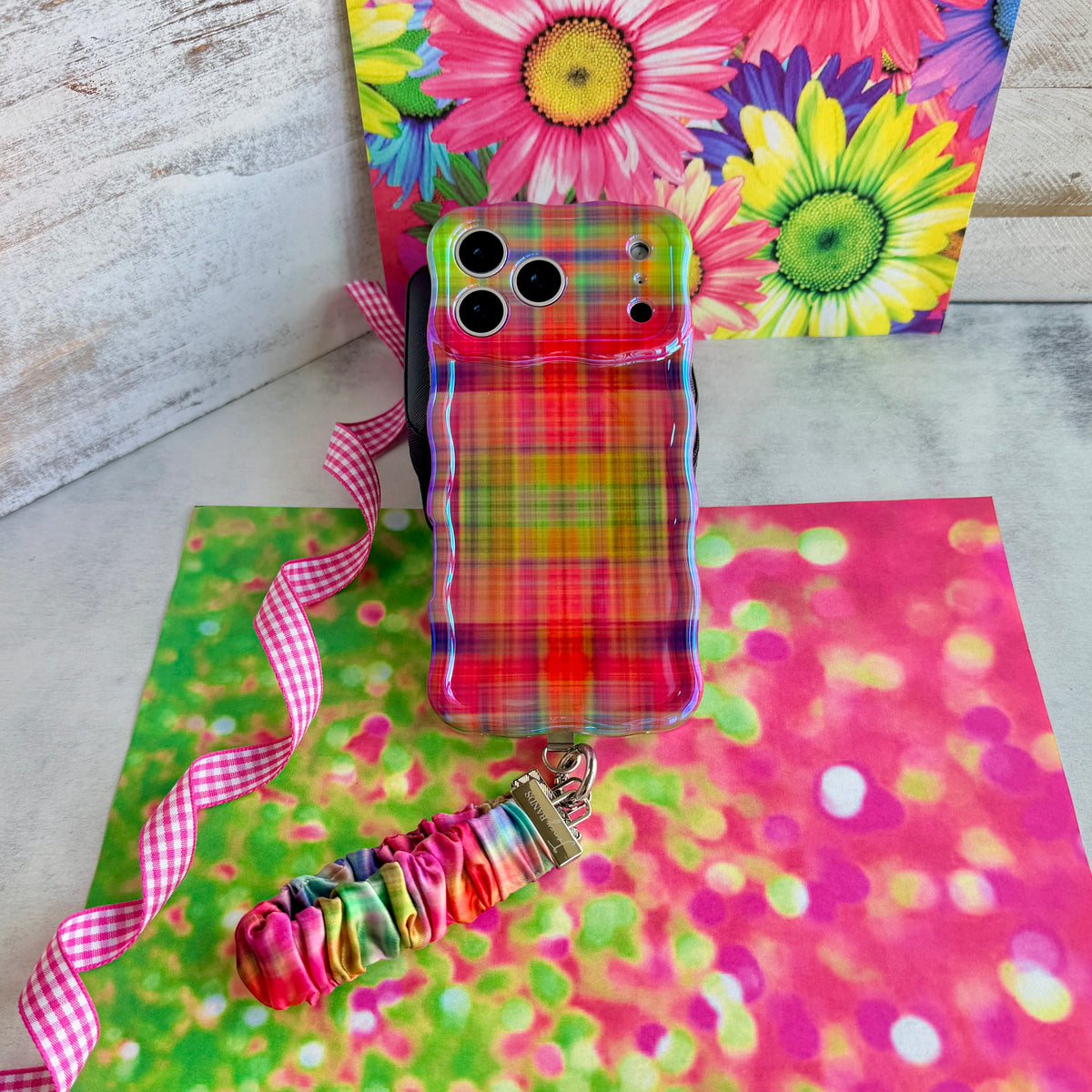 Hazy Tartan scrunchie phone wrist strap laid flat beside the matching blurred rainbow plaid phone case on a bright daisy print background.