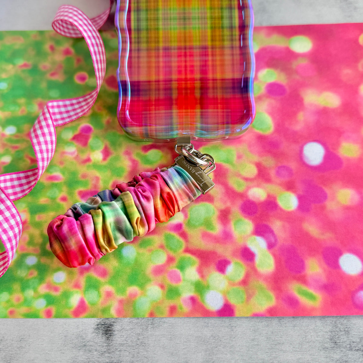 Hazy Tartan Scrunchie Phone Wrist Strap in blurred rainbow plaid, shown with its gingham-print strap on a pink and green confetti backdrop.