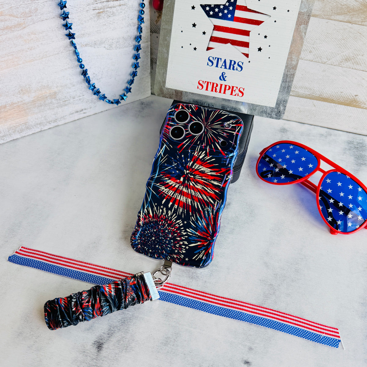 USA Fireworks scrunchie phone wrist strap laid flat with the matching firework phone case, star-spangled sunglasses and a red, white and blue beaded strand.