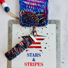 USA Fireworks Scrunchie Phone Wrist Strap in red, white and blue firework print, propped against a Stars and Stripes sign with the matching phone case.