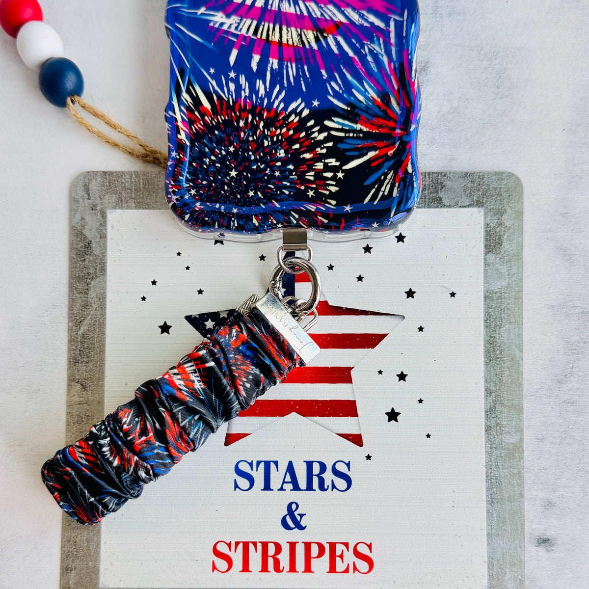 USA Fireworks Scrunchie Phone Wrist Strap in red, white and blue firework print, propped against a Stars and Stripes sign with the matching phone case.