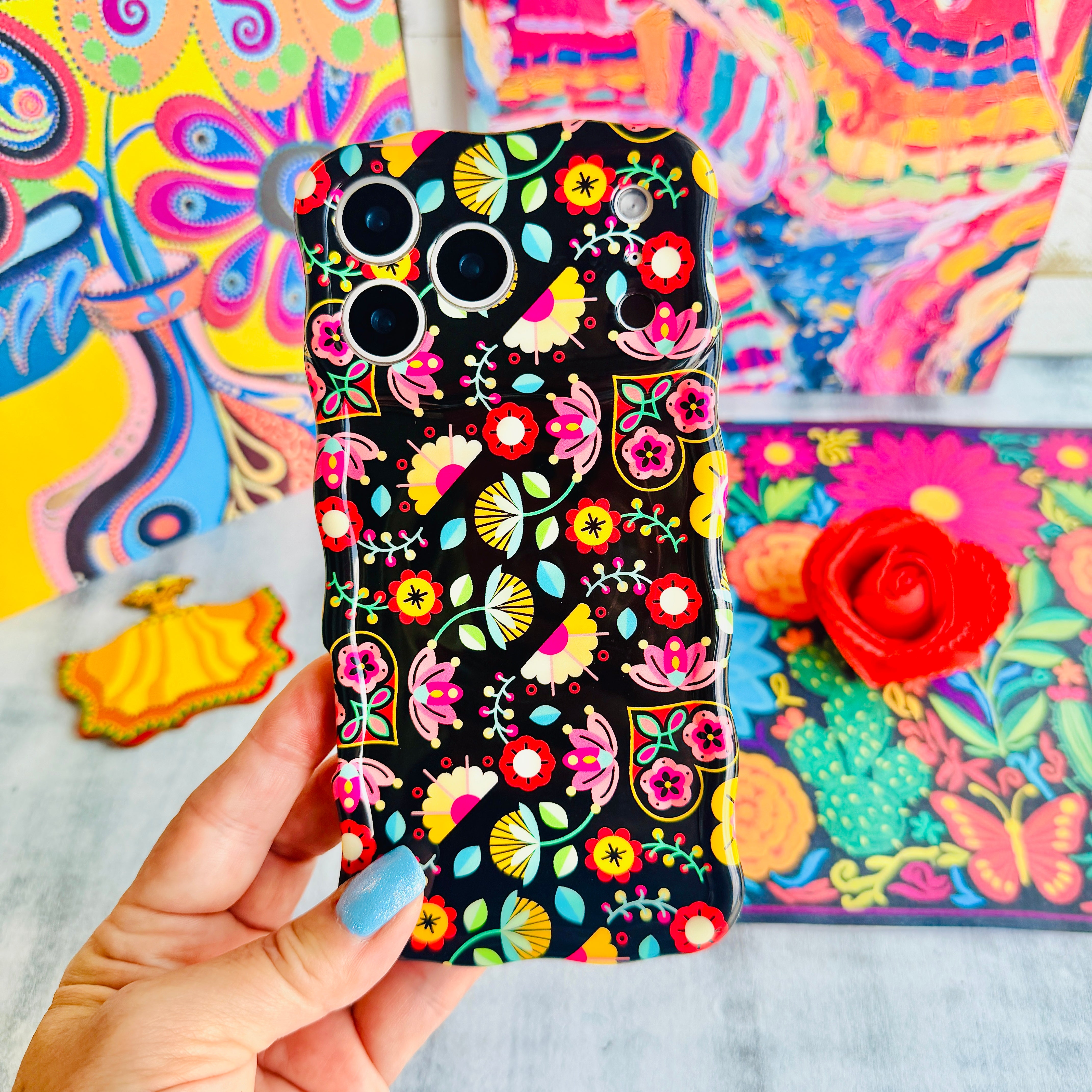 Trendy black wavy iPhone case featuring an intricate folk art botanical pattern of red flowers, magenta details, and yellow fan petals with a high-gloss finish.