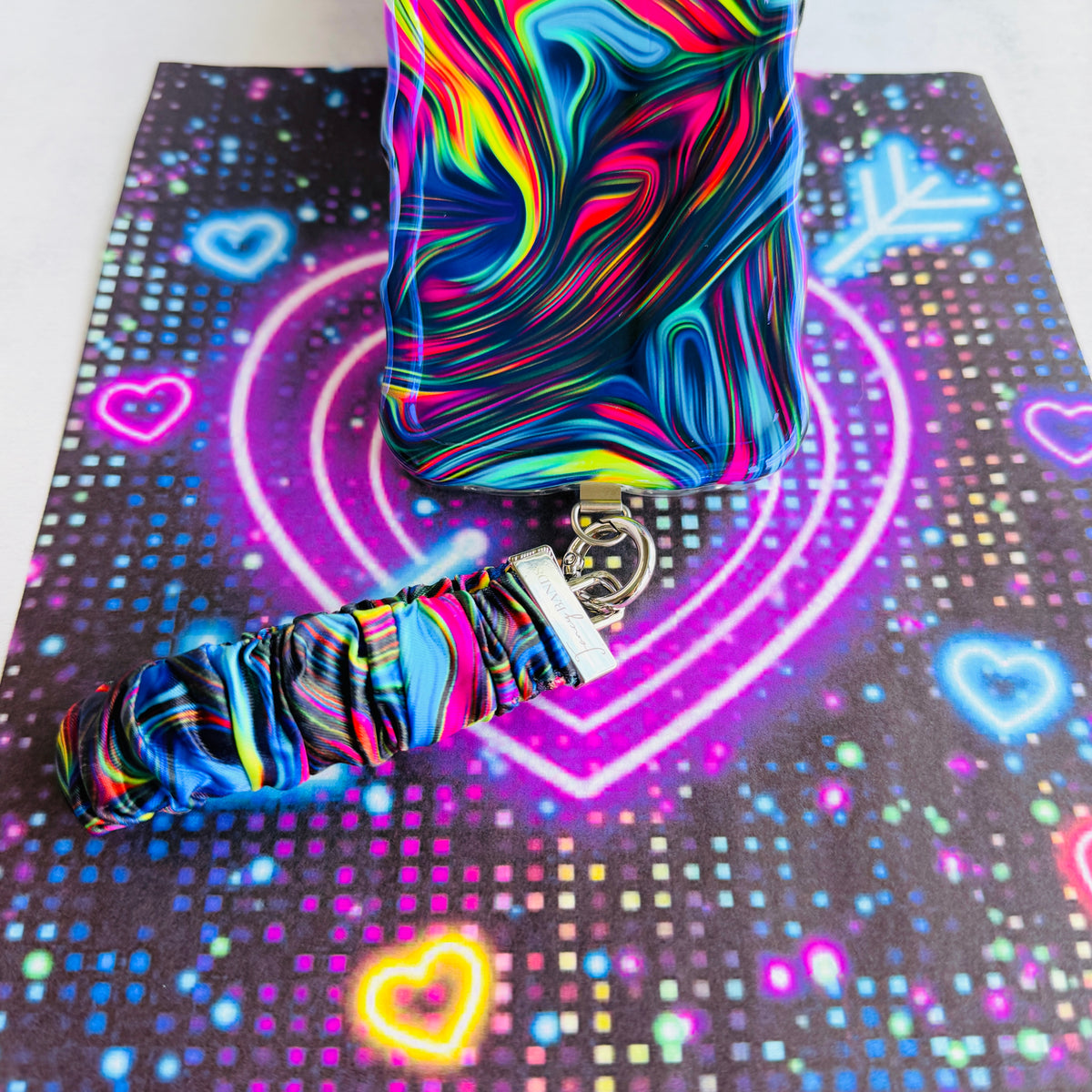 Neon Taffy Scrunchie Phone Wrist Strap in swirled neon marble, standing beside the matching Neon Taffy phone case on a glowing neon heart backdrop.