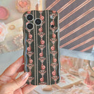 Hand holding the Pretty Little Details black pearl and rose Wavy Luxe iPhone case over a coquette flat lay with a blush compact, pearls and pink roses.