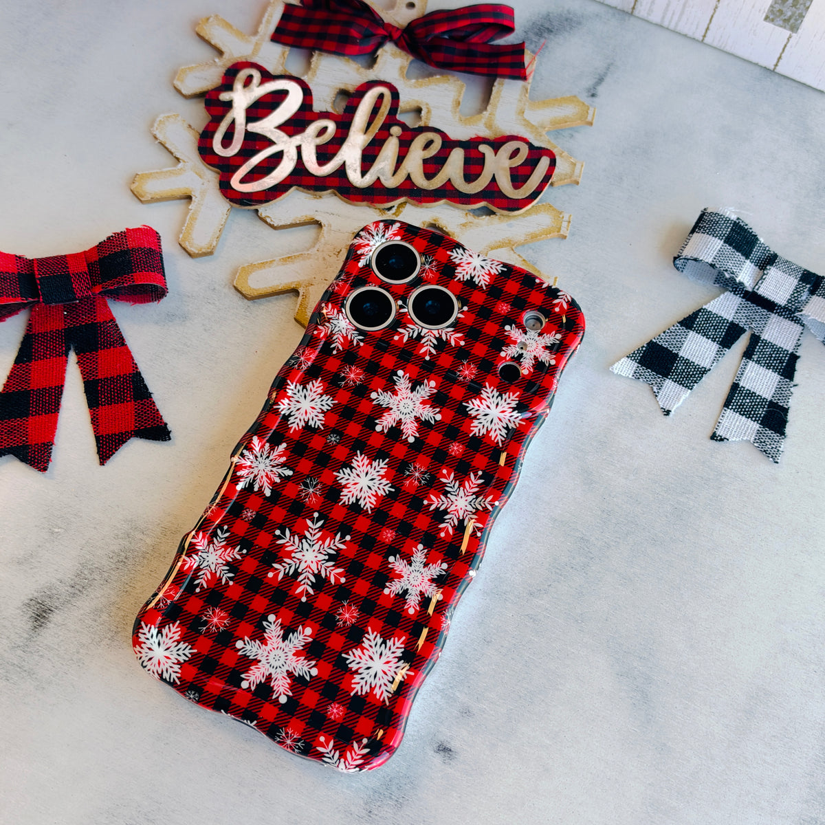 Buffalo Plaid Snowflake Wavy Luxe MagSafe iPhone case laid flat, red and black buffalo plaid with white snowflakes, with a Believe sign and buffalo check bows