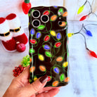 Hand holding the Christmas Lights Wavy Luxe MagSafe iPhone case, glowing multicolor bulbs on black