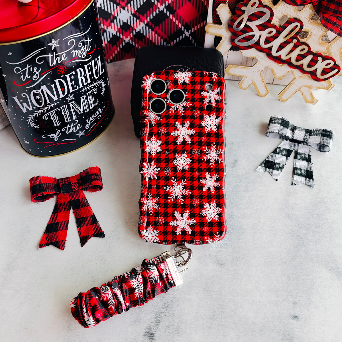 Buffalo Plaid Snowflake iPhone case with the matching scrunchie phone wrist strap attached, laid flat with a Believe sign and buffalo check bows