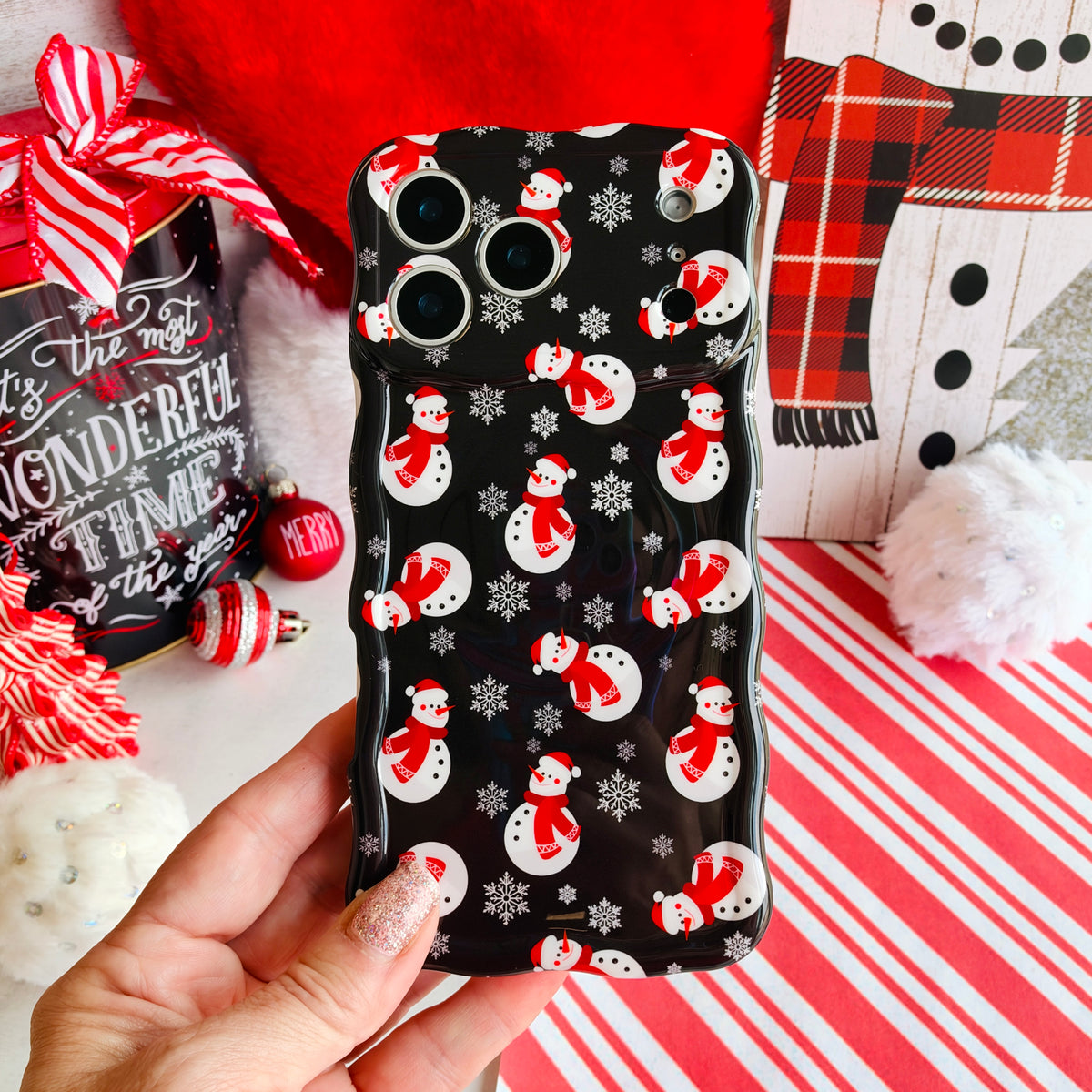 Hand holding the Snowman Wavy Luxe MagSafe iPhone case in black snowman print, on candy cane stripes with a Santa hat and ornaments