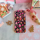 Merry Mugs & Ginger Hugs Wavy Luxe MagSafe iPhone case laid flat, pink gingerbread and cocoa mug print on black, with gingerbread cookies and a gingerbread house