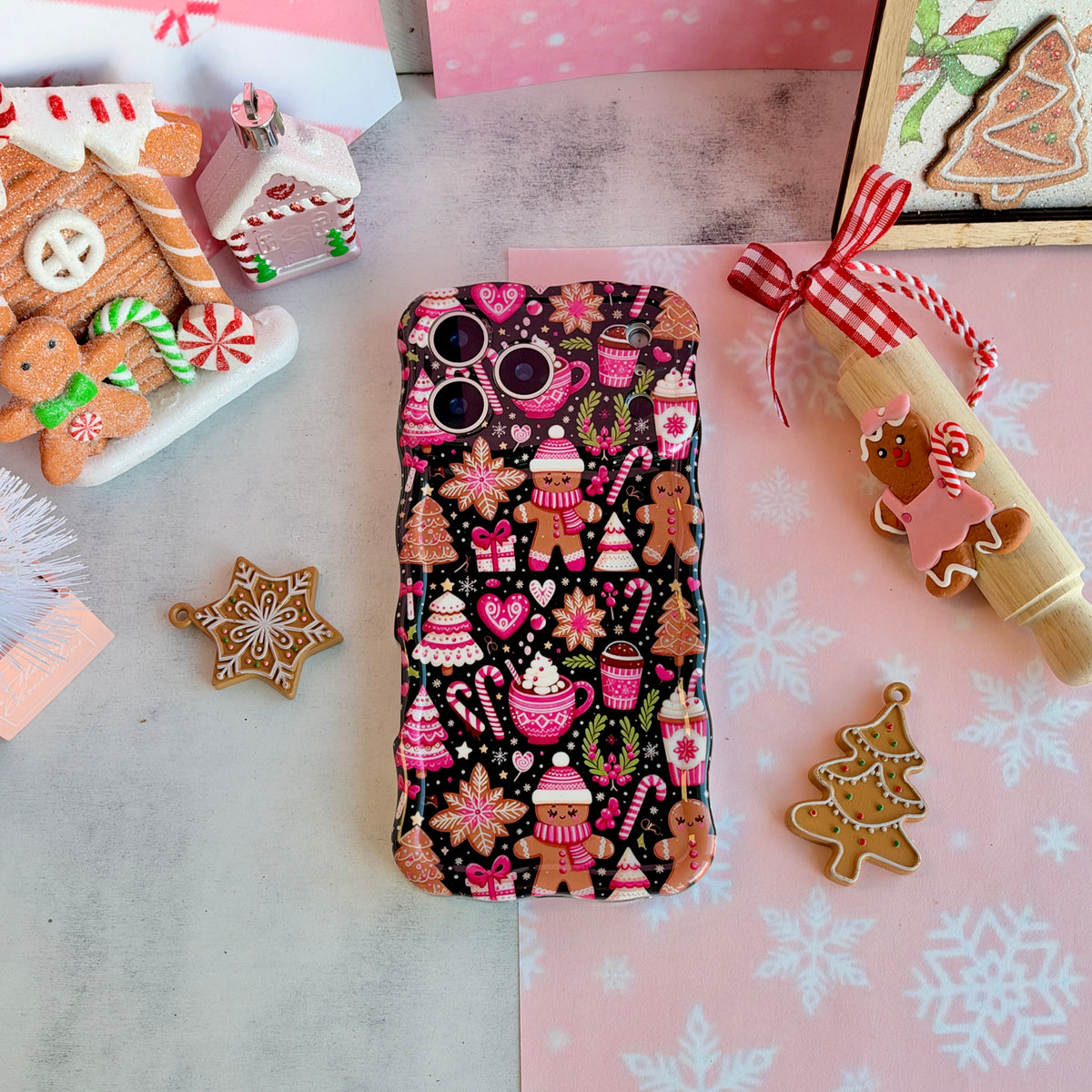 Merry Mugs & Ginger Hugs Wavy Luxe MagSafe iPhone case laid flat, pink gingerbread and cocoa mug print on black, with gingerbread cookies and a gingerbread house