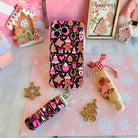 Merry Mugs & Ginger Hugs iPhone case with the matching scrunchie phone wrist strap attached, laid flat with gingerbread cookies and a gingerbread house