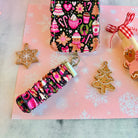 Merry Mugs and Ginger Hugs scrunchie phone wrist strap clipped to the matching gingerbread Wavy Luxe iPhone case, styled with cookies and a gingerbread house