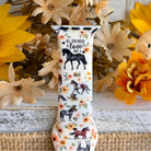 Decorative watch strap with horse design and floral patterns on a wooden surface with flowers.