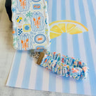 A Coastal Catch scrunchie phone wrist strap featuring a blue tile, orange lobster, and yellow lemon pattern attached to a matching Wavy Luxe phone case on a blue and white striped background.