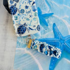 A Blue Lagoon scrunchie phone wrist strap with a blue and white sea turtle and ocean design attached to a matching Wavy Luxe case on a blue starfish background.