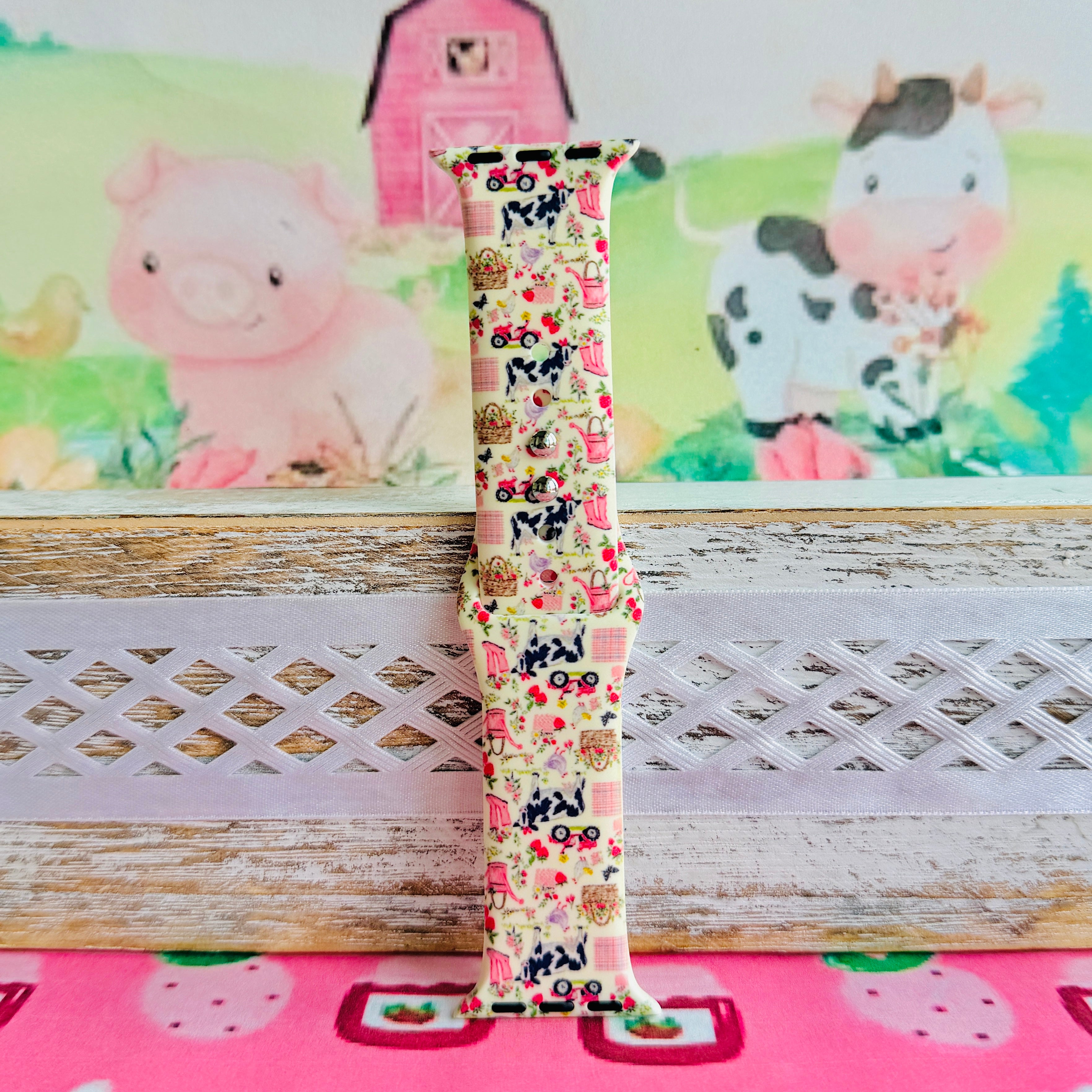 Farm Girl Cutie silicone Apple Watch band with dairy cows, red tractors, chickens, ducks, pink watering cans, strawberries, baskets, and roses on a cream base with pink gingham squares, shown displayed and worn on Apple Watch against a farmhouse country backdrop.