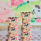Farm Girl Cutie silicone Apple Watch band with dairy cows, red tractors, chickens, ducks, pink watering cans, strawberries, baskets, and roses on a cream base with pink gingham squares, shown displayed and worn on Apple Watch against a farmhouse country backdrop.
